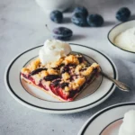 Ein weißer Teller mit einem Stück Zwetschgenkuchen mit Hefeteig und Streuseln, serviert mit Schlagsahne. Im Hintergrund violette Zwetschgen, im Vordergrund ein weiterer Teller.